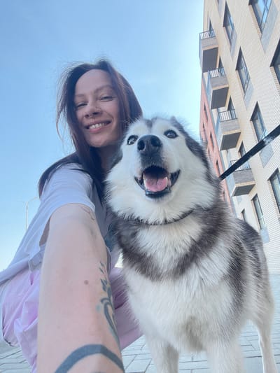 A selfie captures a young woman and a husky-type dog outdoors in Svetloe, Russia. The woman has dark hair and a nose ring, and she is smiling. She is wearing a white t-shirt. Her right arm, which is holding the camera, has tattoos. The husky, with black and white fur, is looking directly at the camera with its mouth open. A black leash is visible above the dog. Behind them is a building with multiple floors and windows, indicating an urban environment. The background also includes a clear blue sky. The setting seems to be a sunny day in the city.