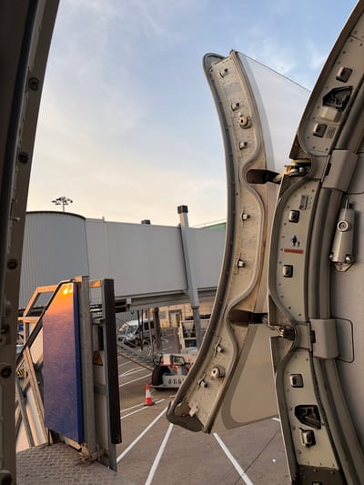 This media file shows an upward-facing perspective of an open aircraft door and its surrounding mechanism, against a bright sky with scattered clouds, suggesting it is daytime. The scene appears to be at an airport or airfield, indicated by the presence of a large building in the background, runway markings, and airport equipment. The sunlight hitting a blue panel suggests it might be late afternoon or early morning.
The primary subjects are the mechanical components of the aircraft door, including hinges, latches, and reinforcing structures, all made of metal and appearing weathered. There are warning labels visible on the interior of the door frame. One label, partially obscured by glare, features a pictogram of a person with an exclamation mark, indicating a potential hazard. Another label, "WARNING", is clearly visible. The overall impression is one of industrial infrastructure and aviation operations.
The angle and the exposure of the image suggest the photograph was taken by someone standing just outside or on the edge of the open door, looking upwards. The lighting suggests a clear, possibly warm day. The location context points to Uttlesford, United Kingdom, which further solidifies the airport setting. No people are explicitly visible in the frame, but the presence of equipment and the open door implies activity related to aircraft maintenance, boarding, or disembarking.