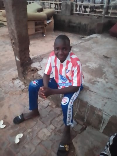 A young boy, possibly of Nigerian descent, sits on a concrete ledge in what appears to be Wuro Shiji, Nigeria. He wears a red and white striped jersey with blue pants that have white accents and the number "32" on the left leg. The boy has his hands clasped in his lap and is looking towards the camera with a slight smile. In the background, there is a large, light-colored sofa or mattress on a wooden structure, suggesting a somewhat informal outdoor living space. The ground is paved with reddish-brown bricks. Two white, curved objects, possibly flower petals or leaves, are on the ground in front of the boy. A dark brown pillar is to the left of the boy, and a metal fence is visible in the far background. The lighting suggests it might be dusk or an overcast day.