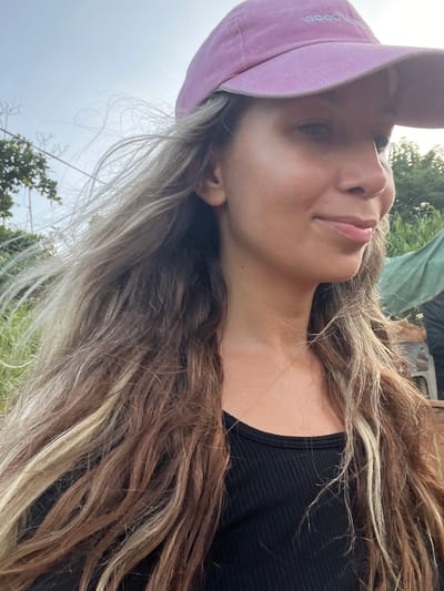 A woman wearing a pink baseball cap that reads "GOOD VIBES" and a black ribbed tank top is shown from the chest up, in a close-up shot. Her long, wavy, ombre-colored hair, with streaks of blonde and brown, is blowing in the wind to her left. She is looking to her right, with a slight smile on her lips. The background is outdoors, with greenery and trees visible, suggesting a rural or natural setting. A hint of a green tarp is visible in the far right of the image. The lighting suggests it is daytime. The location is Talalla, Sri Lanka.