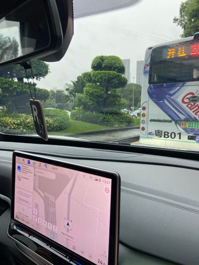 The image, taken from inside a vehicle, shows a traffic scene in Luohu District, China. The driver's side dashboard is visible, featuring a large touchscreen display with a navigation map. The map shows roads, including "Aiguo Road Side," and a blue arrow indicating the vehicle's current position. The exterior view through the windshield includes a bus with Chinese characters and the destination display showing "开往 2," indicating a route. Other cars and lush green trees are also visible in the background, suggesting an urban environment with significant greenery. A novelty air freshener hangs from the rearview mirror. The weather appears overcast.