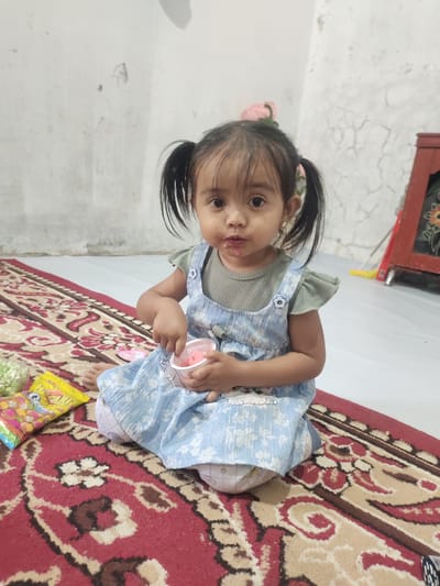 A young girl with dark hair in pigtails sits on a red and cream patterned rug, holding a small pink cup and a spoon. She is wearing a light blue dress with a floral pattern over a green top. The wall behind her is white and textured, with some visible cracks. To her left, on the rug, are a yellow package of snacks and a green bag. A red cabinet with floral details is visible to her right. The scene appears to be indoors, in a residential setting in Cintadamai, Indonesia.