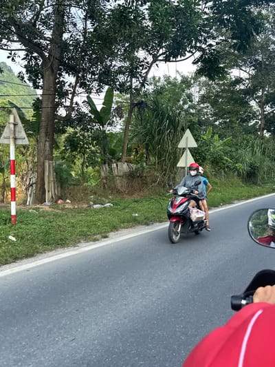 An outdoor, daytime image captures a scene along a paved road, likely taken from a moving vehicle. The focal point is a red and black scooter carrying two individuals.
The driver, an adult, is wearing a grey hoodie, a red helmet with a white stripe, and a white face mask. Behind the driver, a smaller person, possibly a child, is lying horizontally across the back seat of the scooter. This passenger is dressed in a light blue top and shorts, with sandals on their feet, and their head rests near the driver's back. A white plastic bag is visible on the scooter near the child's feet.
The road is dark grey asphalt with a white line marking its edge. To the left, a green roadside verge is abundant with grass and dense vegetation, including tall, leafy trees, bushes, and broad-leafed plants that resemble banana leaves. Some discarded litter is visible on the grass. Two white triangular road signs with black arrow outlines, both pointing to the left, are mounted on poles within the vegetation. Lower down, a red and white striped pole or barrier is partially seen. The background, faintly visible through the trees, suggests a hilly or mountainous landscape.
On the right side of the image, the photographer's perspective is evident, with a rearview mirror showing a reflection of a helmeted person, and a person's arm in a red sleeve holding onto a part of the vehicle. The scene depicts travel on a rural or semi-rural road during clear daylight hours. There is no visible text in the image.