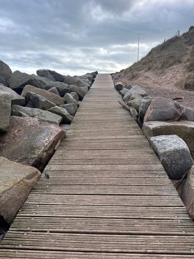 This image depicts a weathered wooden boardwalk leading upwards towards an indistinct horizon, flanked on both sides by large, irregularly shaped grey and reddish-brown rocks serving as a barrier. The scene is located in Montrose, United Kingdom. The boardwalk, constructed from parallel wooden planks secured by visible metal fasteners, occupies the central perspective, narrowing as it recedes. To the right of the boardwalk and beyond the rocks, a sandy dune rises, sparsely covered with dry, light-brown grasses. A single thin, light-colored pole stands vertically on this dune, with a section of a dark fence visible along the dune's crest in the distance. Some driftwood is observed amongst the rocks on the right side. The sky overhead is uniformly overcast with shades of grey, indicating a cloudy day. There are no individuals present or visible actions occurring in the scene.