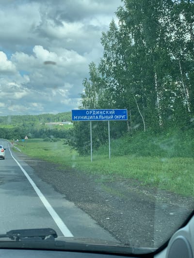 The image shows a blue road sign in Russian, indicating the location "ОРДИНСКИЙ МУНИЦИПАЛЬНЫЙ ОКРУГ" (Ordinsky Municipal District). The sign is located on the side of a road, likely a highway, with a grassy verge and trees in the background. The weather appears overcast with clouds. A car is visible in the distance on the left side of the road. The scene is set in a rural or semi-rural area. There is no visible information in the image that places the scene in the city of Суда, Russia.