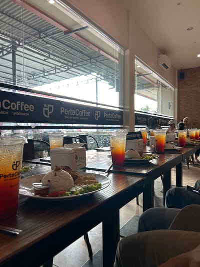The image depicts the interior of a cafe in Meunasahceubrek, Indonesia, with a focus on a long wooden table laden with food and drinks. In the foreground, a plastic cup filled with an amber-colored beverage with a red gradient is prominent. Adjacent to it, a white plate holds a meal consisting of rice, fried chicken, cucumber slices, and a small container of sauce. A white tissue box with "LIVi" branding sits on the table between the drinks and food. Further down the table, multiple other beverages in similar plastic cups, some layered with orange and red hues, are visible, along with plates of food. Several glasses of iced tea and other drinks are present. In the background, through a large window, the exterior of what appears to be an open-air structure with a metal roof is visible. People are seated at the table, with their backs or profiles to the camera, engaged in dining or conversation. The cafe's interior features brick walls on the right side, and a modern air conditioning unit is mounted on the wall above the window. The overall lighting suggests an indoor setting with natural light coming from the window. A banner with "PertaCoffee" branding is displayed above the window.