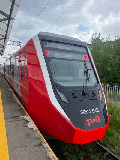 A bright red and white electric train, identified by the number "3C104-045" and the Russian Railways logo "РЖД", is stopped at a platform in the city of Lomonosov, Russia. The train has a sleek, modern design with large tinted windows and red LED marker lights. A yellow safety line is visible on the dark asphalt platform, which appears wet, suggesting recent rain. Overhead, a gray, overcast sky is visible, with visible power lines and a section of a station's canopy. Green foliage and a white metal fence are present in the background. The image is taken at a slightly low angle, emphasizing the train's front and the platform.