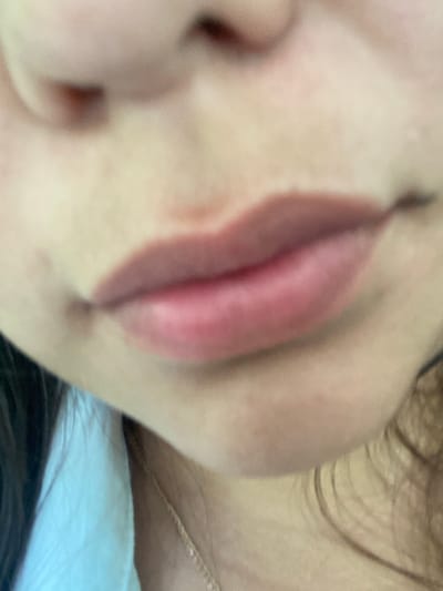 This is a close-up, slightly out-of-focus image of a person's mouth and lower face. The lips are slightly parted and appear to have a pink or reddish tint, possibly from lipstick or natural coloring. There are some freckles or blemishes visible on the skin. The person is wearing a light blue garment, possibly a shirt or blouse, and a thin gold necklace can be seen. The background is blurry and does not provide any clear environmental details. The image does not contain any information about the city of Tyulyugul, Russia.