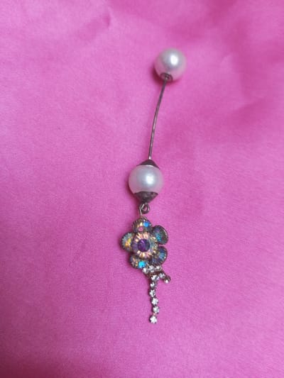 The image displays a close-up view of a decorative jewelry item positioned on a bright pink fabric surface. This scene is situated in Qarabucaq qəsəbə, Azerbaijan. The jewelry piece consists of two white, spherical, pearl-like beads, each approximately 1 cm in diameter, connected by a slender, curved silver-toned metal shaft. A pendant is suspended from the lower pearl bead. This pendant is designed as a flower, approximately 1 cm in width, intricately set with multiple small, iridescent rhinestones exhibiting a spectrum of colors including purple, blue, and gold. Below the floral component, a linear dangle comprising seven small, clear, round rhinestones extends downwards. The metal settings of the pendant appear silver-colored or slightly tarnished. The vibrant pink fabric background exhibits subtle folds and creases. No human subjects, specific actions, or direct interactions are depicted in the frame.