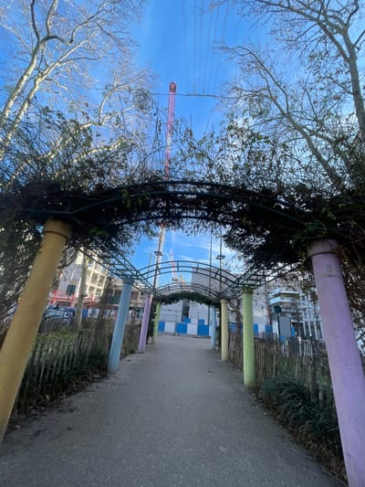 The image displays a paved pedestrian pathway in Gennevilliers, France, viewed from a low angle. The pathway leads through a metal arched trellis structure, which is covered in dark, mostly bare vines with some green leaves. This trellis is supported by multiple cylindrical pillars painted in varying pastel colors, including yellow, light blue, light purple, and light green. Low wooden fences border the pathway on both sides.
Beyond the archway, a construction site is visible, enclosed by blue and white temporary barriers. Within the construction zone, the partially constructed framework of a building and several red and white tower cranes are prominent. In the background to the left, a multi-story building and a dark-colored vehicle are discernible. The upper portion of the scene is dominated by a clear blue sky with minimal white clouds. Bare deciduous trees with intricate branches frame the top edges of the image, with some overhead utility lines visible against the sky. No individuals are present in the visible scene.