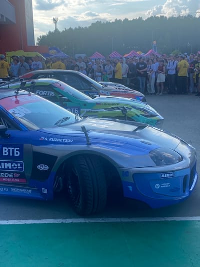 This image depicts an outdoor event with a crowd of people gathered behind a barrier, observing several race cars. Two cars are prominently featured in the foreground, with a third car visible behind them. The race cars are highly decorated with sponsor logos and racing numbers. The blue car in the closest foreground has the number "36" visible on its windshield and displays logos such as "ВТБ," "AIMOL," and "RDSTV.RU." The car behind it is green and has logos like "FORTIS" and "RTE." The background shows trees and a cloudy sky. A crowd of spectators, dressed casually, are standing and watching the scene. The event appears to be a race or car show, possibly taking place in a publicly accessible area. The text "Берёзовка, Russia" is provided as a location, but there are no specific visual cues within the image that definitively identify it as this location. The overall atmosphere suggests a daytime event with good weather.