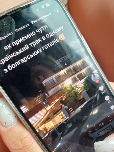 The image is a close-up shot of a smartphone displaying a video. The phone screen shows text in Ukrainian, which translates to "It's amazing to hear about a Ukrainian trek in one of the Bulgarian hotels." The video appears to be a travel vlog or a similar content, featuring a modern building with balconies, illuminated at night. A car is visible in the foreground, with a license plate that reads "EC 0727NG". There are also social media engagement icons, including a heart with "9477" likes and a speech bubble with "155" comments. The phone is held by a person whose fingernails, adorned with rhinestones, are visible in the lower left and right corners of the image. The background is out of focus, suggesting an indoor or outdoor setting outside the frame of the phone. There is no specific indication of the city of Dropla, Bulgaria within the visible content of the image.