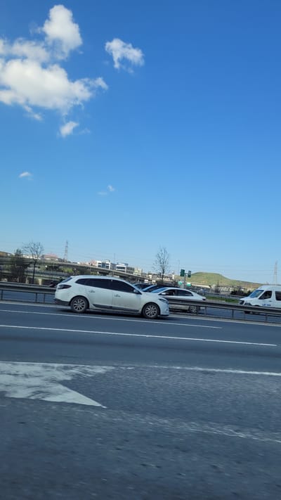 The image shows a section of a highway in the city of Bağcılar, Turkey. The sky is blue with scattered white clouds. In the center of the image are several white vehicles travelling alongside each other. Utility poles line the edge of the road. A grassy hill is faintly visible in the background.