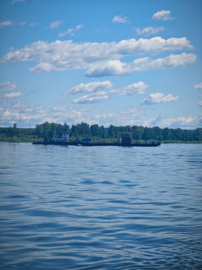 A wide, blue river with gentle ripples dominates the foreground. In the middle ground, a long, dark-colored barge is being pushed by a white and black tugboat. The tugboat has a cabin structure with windows. The barge appears to be carrying some sort of industrial equipment. A treeline of green trees forms the background, with a bright blue sky filled with scattered white clouds above. The scene suggests a sunny day in a rural or semi-rural location, possibly on the outskirts of Krutoi Log, Russia, given the industrial nature of the vessel and the natural landscape.