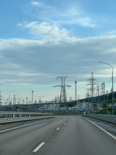The image shows a road running alongside a high-voltage power line infrastructure. The road is made of asphalt and has a white lane marking. There is a gray safety rail. On the left is the power infrastructure and a cloudy sky. The scene is taken from a high angle. Based on the available information, it's impossible to verify if the location is indeed in the city of Inkerman, Ukraine.