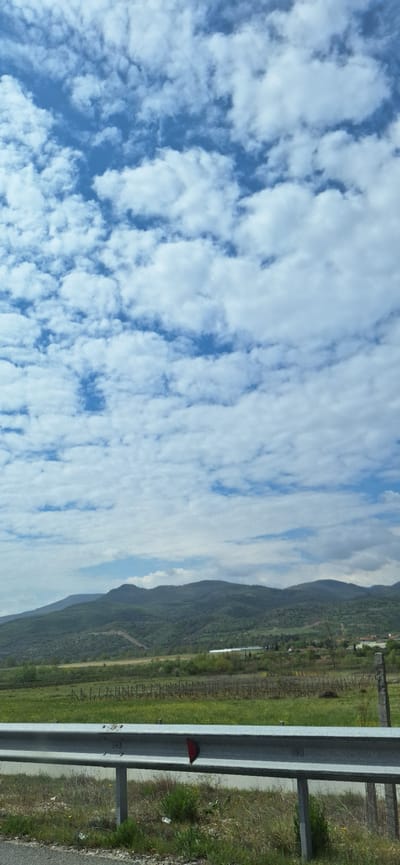 The scene depicts a roadside view in what is suggested to be near Valkovo, Bulgaria. The sky is mostly blue, filled with patterned clouds. Adjacent is a mountainous landscape with greenery. At the edge of the frame are metal guardrails, with vegetation growing around them. In the distance, there appears to be a vineyard or cultivated land.