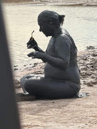 A person covered in dark mud is sitting on the shore of a body of water. The person is facing left and holding a small yellow object in their right hand. The water behind the person is calm with ripples reflecting the light. The shore is made of textured, light brown dirt and rocks. A dark, blurry foreground element is present on the left side of the image. The overall scene appears to be an outdoor natural setting.