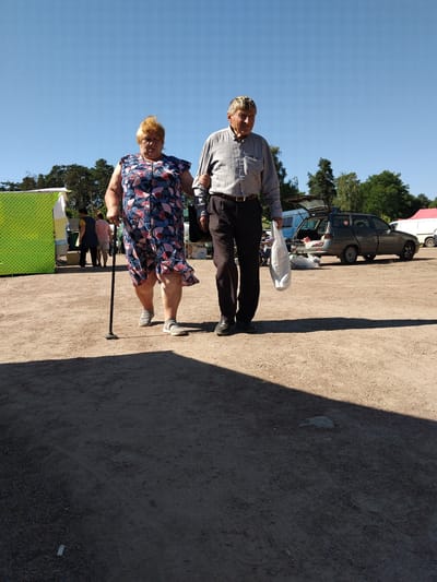An older woman with blonde hair and a colorful patterned dress is walking with a cane and an older man in a gray collared shirt and dark pants. They are walking on a dirt path with a sparse scattering of gravel. Behind them, there are several stalls with green and yellow coverings, and further back, a dark-colored station wagon with its trunk open, parked next to a white van. The sky is clear and blue, and trees line the horizon. The scene appears to be an outdoor market. The image was taken on June 9, 2024, in Irshansk, Ukraine.