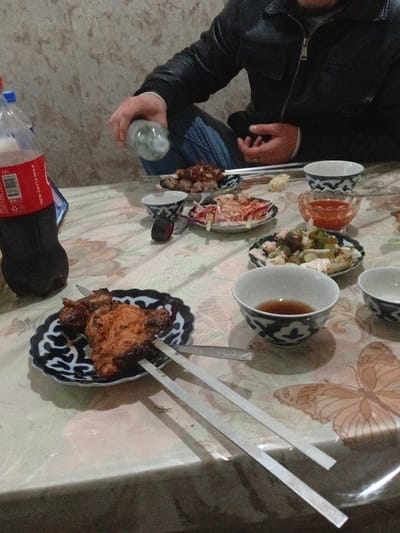 A table laden with food, including meat skewers on patterned plates, salad, and small bowls containing liquid. A man in a black jacket is captured pouring from a bottle. A bottle of Coca-Cola is visible on the left. The tablecloth features a floral and butterfly design. The scene suggests a casual meal setting.