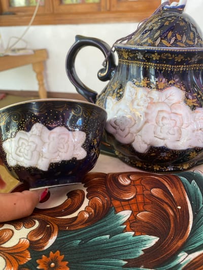 A close-up, eye-level shot focuses on a dark blue porcelain teapot with a lid and a matching cup, both adorned with ornate gold floral patterns and raised white floral designs. The teapot has a dark handle and a decorative lid with a small string attachment. A hand with pink nail polish is visible holding the cup, its fingertip touching the base. The objects rest on a patterned fabric with swirling designs in brown, orange, and teal. In the background, a wooden table with legs is partially visible, along with a window with a wooden frame. The scene appears to be indoors.