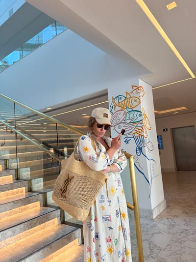 In the city of Pyrgos Community, Cyprus, a woman stands on a marble-tiled floor next to a polished brass handrail of a well-lit staircase. She wears a white, calf-length dress adorned with a colorful pattern of marine life and sun motifs, a beige baseball cap with a logo, and sunglasses. A large, woven straw tote bag with a gold logo hangs from her left shoulder. She holds a smartphone in her right hand, appearing to text. Behind her, a white pillar is decorated with a vibrant mural of stylized fish. The staircase features marble treads and risers, with a glass railing, and is illuminated by embedded lights. An elevator is visible in the background. The overall environment suggests a modern indoor space, possibly a hotel or public building.
