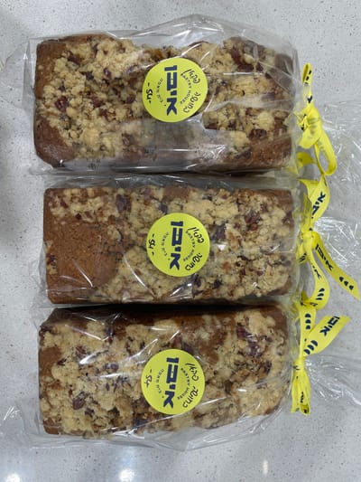 Three rectangular loaves of baked goods, each individually wrapped in clear plastic and positioned vertically in a slight stagger, are displayed on a light-colored countertop. Each loaf features a light brown cake base topped with a generous crumble containing visible fragments of nuts. A circular, bright yellow label is affixed to the plastic wrapping of each loaf. The label displays the text "איקא Pastry House" in both Hebrew and English, along with "קונדיטוריה" (Confectionery/Bakery) in Hebrew, and a price, visible as "54-" on at least one label, indicating 54 Israeli New Shekels. Each wrapped loaf is also secured with a bright yellow ribbon that has the Hebrew name "איקא" (Ika) printed repeatedly in dark blue. The immediate environment is a flat, light grey or off-white surface, consistent with a countertop, likely in a commercial establishment or private residence in Nes Ziona, Israel, given the Hebrew script and implied currency. No individuals are present in the image.