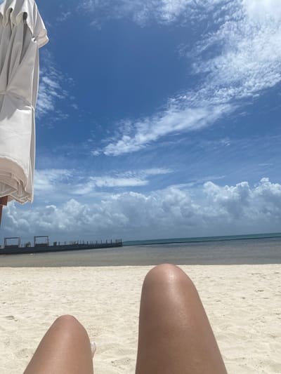 The image depicts a daytime scene at a beach in Marina Maroma, Mexico. The sky, occupying a large portion of the view, is predominantly blue with varying cloud formations. Some clouds are light and whispy, while others are larger and more cumulus-like. The beach itself is sandy and stretches towards the horizon. Parallel to the waterline are dark, evenly spaced structures. To the lower left, a white beach umbrella is partially visible. In the lower right are a person's thighs resting on the sand. One toe is visible.