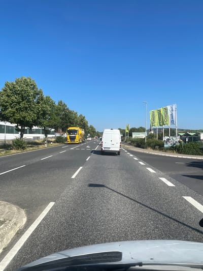 The image depicts a sunny daytime scene on a multi-lane road in Wartenberg, Germany. The road has clear white lane markings and asphalt paving. On the left side of the road, there are trees lining the pavement in front of a low-rise industrial building with white walls and a dark roof. Further down the road on the left, a bright yellow tanker truck is visible. In the center of the road, a white panel van is driving away from the viewer. On the right side of the road, several large flags with green and yellow lettering are displayed. The sky is clear and blue. The image appears to be taken from inside a vehicle, looking forward.