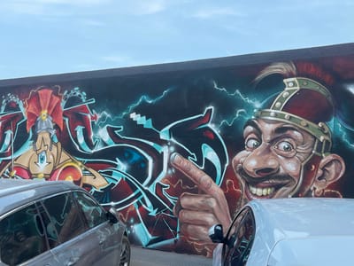 The image captures a section of a vibrant graffiti mural on a dark-colored wall in Cartagena, Spain, beneath a pale blue sky. The mural depicts two caricatured figures. On the left, a stylized figure wears a silver helmet with a red plume and stands behind a geometric design with red and blue accents, reminiscent of lightning. On the right, a large, close-up portrayal of a man's face with wide, bulging eyes and a broad, toothy grin dominates the scene. He wears a similar helmet with green accents. His right hand, with a pointed finger, is prominently displayed in the center of the mural, seemingly interacting with the lightning motif. In the foreground, two cars are parked, a silver SUV on the left and a white vehicle on the right, partially obscuring the lower portion of the mural. The urban setting is suggested by the wall and the parked vehicles.