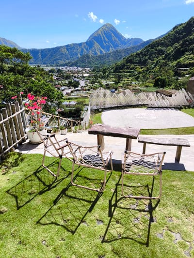 The photograph shows a scenic view in Sembalun Bumbung, Indonesia, featuring a mountain backdrop under a clear, blue sky with scattered clouds. In the foreground, there is a seating area with three tan-colored chairs and a rustic wooden table and bench set on a patch of green grass. A bamboo fence and potted plants with pink flowers border the seating area. Below the seating area, a white picket fence encloses a gravel area with circular patterns etched into the ground. Beyond the fence, a valley stretches out, dotted with buildings and greenery, forming part of the landscape of Sembalun Bumbung. The scene appears to be taken on a bright, sunny day.