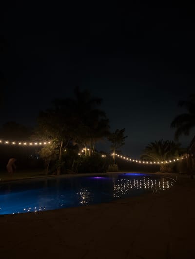 The image displays an outdoor scene at night, characterized by a dark, clear sky and ambient lighting. Along the right edge, a body of water, likely a swimming pool, stretches from the bottom right corner towards the top. The water is a deep blue, reflecting numerous small, bright points of light from above.
String lights, adorned with warm, glowing bulbs, are suspended across the mid-ground of the scene, creating a horizontal line of illumination. These lights are strung amidst the silhouettes of lush trees and palm fronds, which are dark against the night sky and occupy the left and central parts of the frame. The reflections of these string lights are clearly visible on the surface of the pool, adding to the sparkling effect. The pool's edge or surrounding ground is a dark, textured surface, possibly stone or concrete, visible on the right and bottom right.
In the lower-middle section, beneath the string lights and partially obscured by foliage, a faint, light-colored, indistinct shape is visible on the ground, potentially a person. The setting evokes a sense of tranquility and relaxation, possibly a private residence or resort in a tropical climate. The time of day is clearly night, with no visible weather events. No text is visible in the image. The presence of palm trees and general ambiance is consistent with the provided location context of Santa Ana Yancuitlalpan, Mexico.