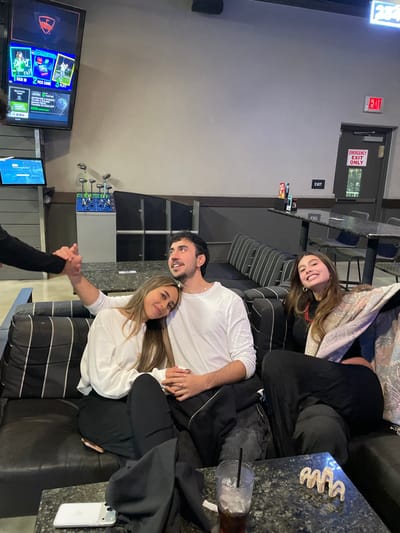 Three people are relaxing on a gray striped sofa. A smiling young woman is seated at the top, while a man in a white shirt and another woman in a white shirt are seated at the bottom, holding hands. A table beside the sofa holds a clear cup with a straw and a white phone. To the left, a television displays a golf game, and golf clubs are mounted on the wall.