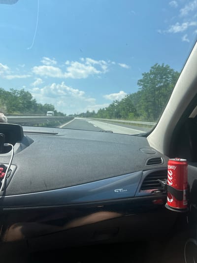 The image shows the interior of a car, looking out through the windshield. The view outside the car is a bright, sunny day with a blue sky and scattered white clouds. Green trees line a highway, suggesting the car is in motion.
Inside the car, a red Coca-Cola can is attached to an air vent on the dashboard with a black holder. The can has the Coca-Cola logo visible. To the right of the air vent, there is a dark, shiny trim piece with "GT line" written on it in silver lettering. A black dashboard is visible in the foreground, with some scratches on its surface. A phone mount with a white charging cable is also present.
The image is taken from the perspective of a passenger or driver. The lighting suggests it is daytime, likely mid-morning or afternoon, given the bright sunshine. The scene appears to be a road trip through a wooded area, consistent with the provided location context of Rajana, Bulgaria. There are no people visible in the image. The overall impression is one of a casual drive on a pleasant day.