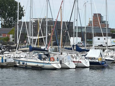 The image shows a view of a marina in Wismar, Germany, with several sailboats docked. The foreground consists of rippling grey water. In the mid-ground, multiple white sailboats with blue trim are moored alongside a wooden pier. Some of the boats have their sails furled and covered with white or blue tarps. One sailboat in the foreground has an orange life preserver attached to its stern. In the background, modern buildings with dark wooden facades and large windows are visible, along with a tall brick building with multiple floors and a stepped roofline. A white camper van with "www.filmbu..de" printed on it is parked behind the boats. The sky is overcast and grey. There are no visible interactions between people, and the scene is static.