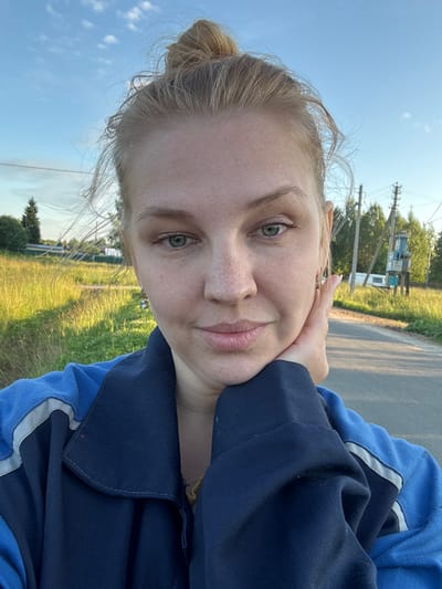 A young woman with blonde hair in a bun is taking a selfie outdoors. She is wearing a dark blue jacket with blue and gray accents. Her left hand is resting on her chin, and she is wearing a small, light blue earring. The background shows a grassy field bathed in sunlight, a paved road, trees, and utility poles under a clear blue sky with some wispy clouds. The location is identified as Dunilovo, Russia.