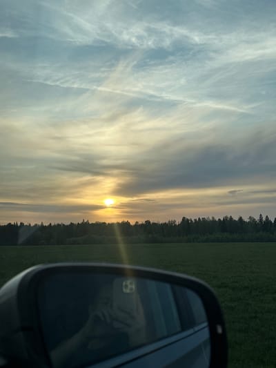 The image depicts a landscape during either sunrise or sunset, viewed partially through the side mirror of a car. The sky is filled with layered clouds, with the sun appearing low on the horizon, emitting a golden glow. To the right, a field or meadow extends towards a line of trees. The car's side mirror is visible in the foreground, reflecting a person holding a phone. The location cannot be definitively determined as Postol, Russia, from the visual information alone.
