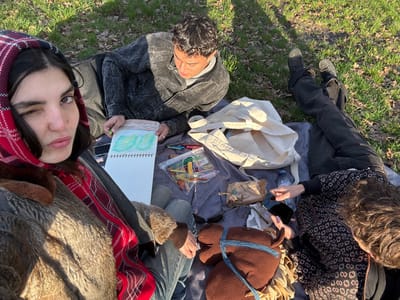 Three individuals are depicted outdoors, situated on a dark blanket spread over a grassy area in the city of Saint-Mandé, France. The female in the immediate foreground, with dark hair, wears a red plaid hooded garment over a fur-lined jacket and jeans, and is captured in the act of winking at the viewer. To her right, a male with short dark hair, dressed in a dark grey jacket and dark pants, lies prone, actively drawing in a white spiral-bound sketchbook with a green implement; the drawing within the sketchbook illustrates green, undulating hills. Further right, a third individual with curly brown hair, wearing a dark patterned long-sleeved top and black pants, lies on their back/side, manipulating a black smartphone with their right hand.
Items arrayed on the blanket include a light-colored canvas tote bag, a plastic water bottle with a blue cap, a small brown paper bag, dark sunglasses, a transparent plastic bag containing various colored drawing implements, and a brown plush horse-head toy featuring a blue strap and a light-colored mane. The surrounding environment consists of green grass with sparse patches of dried leaves, under bright daylight conditions which cast distinct shadows.