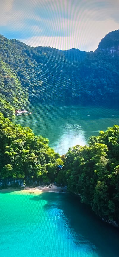 The image displays a serene tropical bay, characterized by lush green, densely forested karst islands rising from the turquoise water. A small, sandy beach is visible on the left side, nestled between the rocky shorelines. A traditional boat with a red roof is moored in the bay. The water is clear and reflects the bright sunlight, creating shimmering patterns. In the distance, several smaller boats are scattered across the water. The sky above is a pale blue with some wispy clouds, and the sun appears to be setting or rising, casting a warm glow. The scene evokes a sense of natural beauty and tranquility. This image is not of Lippstadt, Germany.