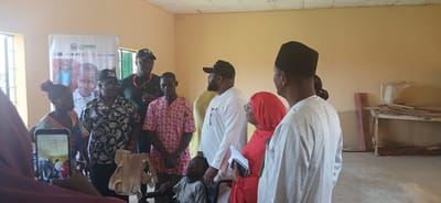In Sabon Gari South, Nigeria, a group of individuals, predominantly adults with one child in a wheelchair, are gathered indoors. The room appears to be a community hall or classroom, with a banner bearing the ROOSC logo and images of children displayed on the wall. The participants are diverse in their attire, ranging from casual to traditional Nigerian clothing, and some are wearing headwear. A woman in the foreground is holding a smartphone, seemingly recording the event. Several people are looking towards the center of the gathering, where a child sits in a wheelchair and another individual is holding a wooden object, possibly a toy or a prop. A table with various items and construction materials is visible in the background, suggesting the room may also serve as a workshop or storage area. The atmosphere seems to be one of an organized event or meeting, with a sense of purpose and community engagement.