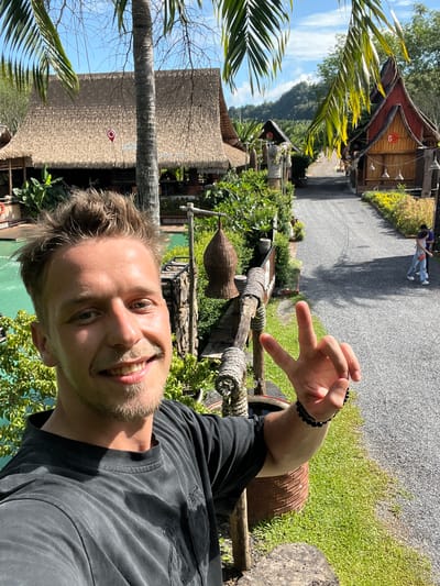 In Tambon Pa Kae Bo Hin, Thailand, a young man with blond hair and a mustache smiles at the camera, making a peace sign with his right hand. He wears a black t-shirt and a black beaded bracelet. In the background, a turquoise swimming pool is visible, surrounded by lush green foliage. Several thatched-roof buildings with wooden accents are scattered throughout the landscape, some featuring decorative elements. A gravel path winds through the scene, with two individuals walking in the distance. The sky is clear and blue with some clouds, and mountains rise in the background.