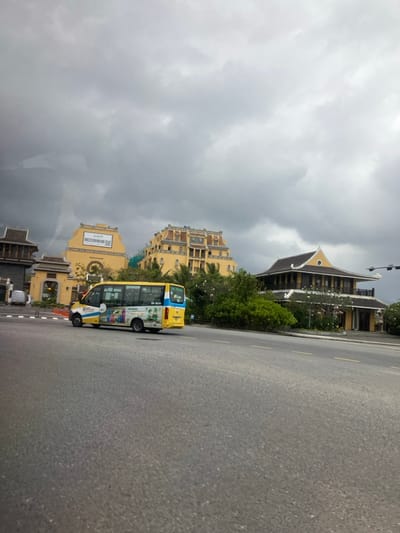 The image depicts an outdoor scene in Hải Vân Ward, Vietnam, on an overcast day. A wide asphalt road occupies the foreground and midground. On this road, a yellow and blue minibus with advertising on its side, including the text "LÀNG VIỆT NAM," is in motion, traveling from left to right across the frame.
In the background, a series of yellow-colored buildings with distinct architectural styles are visible under a heavy, grey, overcast sky. From left to right:
1. A partial view of a building with a dark, multi-tiered roof and light-colored walls.
2. A prominent, yellow, gate-like structure featuring an archway. A rectangular sign on its upper facade displays a phone number "0909 09 09 09" and a QR code.
3. A large, multi-story yellow building with a complex, dark-tiled roof system. Green construction netting is visible on its left side, suggesting ongoing construction or renovation.
4. Further to the right, another distinct building with yellow walls and a pronounced, dark, multi-tiered traditional Vietnamese-style roof stands amidst green foliage.
5. On the far right, a traffic light pole is partially visible.
No individuals are clearly discernible. The environment features maintained landscaping with green trees and shrubs between the road and the buildings. The overall scene suggests a developed urban or resort area.