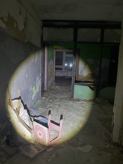 In an abandoned building, possibly a former kindergarten or children's facility, in the village of Yekaterinovka, Russia, a flashlight illuminates a debris-strewn hallway. The walls are peeling, revealing layers of paint in various states of decay, with a prominent green section on the right side bearing faint, abstract drawings. A doorway ahead leads to another room with windows. In the foreground, a pink wooden child's chair, overturned and damaged, rests amidst scattered debris, including sacks and wooden planks. The floor is cracked and littered with rubble. The scene suggests neglect and the passage of time, typical of abandoned structures.
