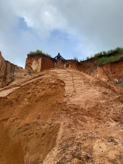A person wearing a dark blue shirt and dark pants is sitting on the edge of a steep, red earth cliff with their legs extended forward and arms out to the sides. The person is positioned in the center of the frame, against a cloudy sky. The cliff face is eroded, with visible layers of soil and some patches of green grass growing on the top edge. The foreground shows a steep slope of loose earth and small rocks. The environment appears to be a natural geological formation, possibly a quarry or a natural erosion site, located in Nguzu Edda, Nigeria.