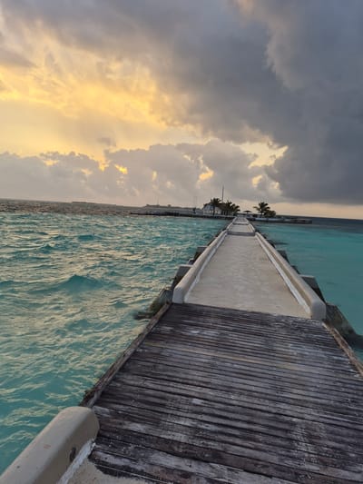 Captured from a low-angle, an expansive view unfolds, displaying a weathered wooden pier stretching into a turquoise sea beneath a sky of dramatic clouds. To the left, the sky transitions from heavy, dark storm clouds to patches of vibrant yellow sunlight breaking through. The sea, reflecting the sky's hues, ripples with gentle waves. The pier, constructed with a central concrete pathway flanked by wooden planks, shows signs of age and wear. In the distance, palm trees and structures dot the horizon, indicating an island locale. This is likely Bodufolhudhoo, Maldives, known for its island piers and surrounding sea. No people are visible in the frame. The dominant action is the natural movement of the sea and the atmospheric interplay of light and clouds.