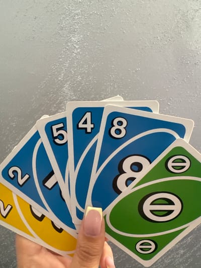 A hand holds a stack of Uno cards, displaying blue cards with the numbers 2, 5, 4, and 8, a green "skip" card, and yellow cards with the number 7. The background is a textured, light gray surface. The scene appears indoors with artificial lighting. There is no indication of location in the city of Glojene, Bulgaria.