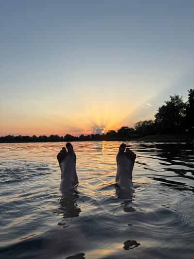 This media file features a person's feet submerged in calm water, with the setting sun visible on the horizon. The sun's rays are dramatically breaking through clouds, creating a beautiful spectacle of light. The water reflects the warm hues of the sunset, casting a golden glow on its surface. The surrounding area appears to be a natural landscape, possibly a lake or river, with a line of trees silhouetted against the vibrant sky. The overall mood is serene and tranquil, evoking a sense of peace and enjoyment. The time of day is clearly sunset, indicated by the low angle of the sun and the warm, soft light. There is no visible text in the image. The location context of Schweinfurt, Germany, suggests this could be a local swimming spot or a scenic overlook.