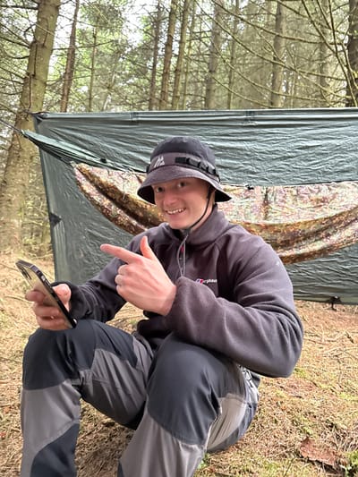 A young man is sitting in a forest, likely Macclesfield Forest in the United Kingdom, at latitude 53.26 and longitude -1.991. He is wearing a grey fleece jacket with the word "Berghaus" on it, a black bucket hat, and grey and black hiking pants. He is sitting on the ground with a large green tarp behind him. The tarp is likely a camping shelter, with a hammock visible underneath it. He is holding a smartphone in his left hand and pointing to the left with his right hand. He is looking directly at the camera with a slight smile. There are trees in the background. The ground is covered in dry leaves and twigs. The weather appears cloudy.