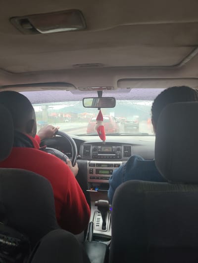The image shows the interior of a car, viewed from the passenger side looking towards the driver. A person wearing a red hoodie is driving, with their hands on the steering wheel. A passenger wearing a blue denim jacket is seated in the front passenger seat, their head and shoulders visible. The dashboard of the car, including the radio and gear shift, is also visible. Outside the car, it appears to be daytime, with overcast or rainy weather, as indicated by the streaks on the windshield. The view through the windshield shows traffic and buildings, suggesting an urban or semi-urban environment. A small, festive-looking decoration, possibly an elf, is hanging from the rearview mirror. The overall scene depicts a moment of travel in potentially adverse weather conditions.