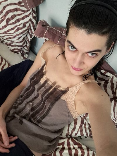 An individual is depicted in an indoor setting in Châtelaillon-Plage, France. The person, appearing female, is positioned on their back, looking directly at an overhead camera, indicating a self-portrait. Dark hair is pulled back by a black headband, with some damp-looking strands on the forehead and temples. Prominent dark eyebrows frame light-colored eyes, and pink lip color is present. The individual wears a sleeveless top featuring a light peach-toned, textured upper section and a grayish-brown lower portion with dark, wavy horizontal stripes. The visible left hand rests on dark blue fabric near the waist. The background consists primarily of bedding or cushions displaying a brown and white zebra-stripe pattern, along with a plain brown patterned cushion and a light blue wall or headboard surface behind the head. Small dark epidermal marks are visible on the right arm and upper chest.
