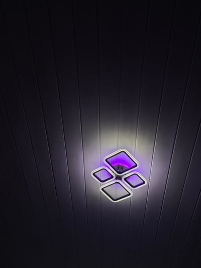 A modern, multi-faceted ceiling light fixture with purple and white illumination is centrally positioned against a dark, wood-paneled ceiling. The light fixture, composed of four interconnected square frames, casts a glow that highlights the wood grain of the ceiling. The overall scene is dark, suggesting it is nighttime or indoors with low ambient light. The location is specified as Южное Снт, Russia, though no specific city is mentioned.
